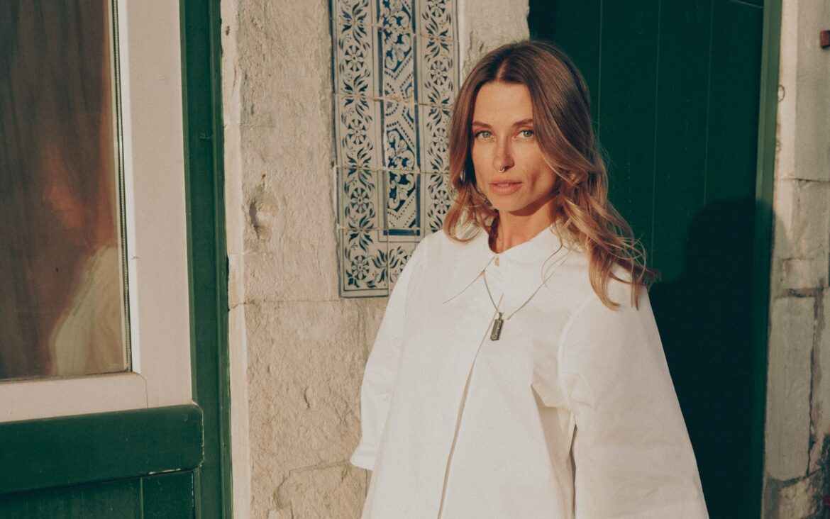 Kate Zubok in a white outfit beside a green doorway, in an artist-supplied press portrait.