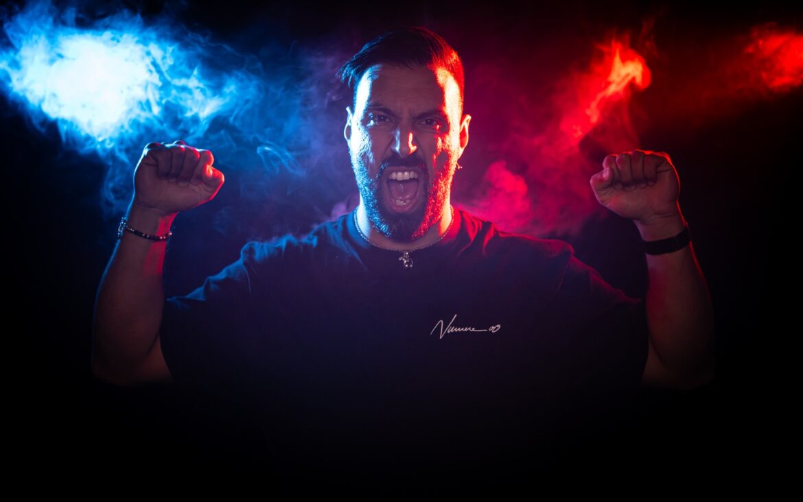 Massimo Conte in a blue-and-red-lit portrait with both arms raised.
